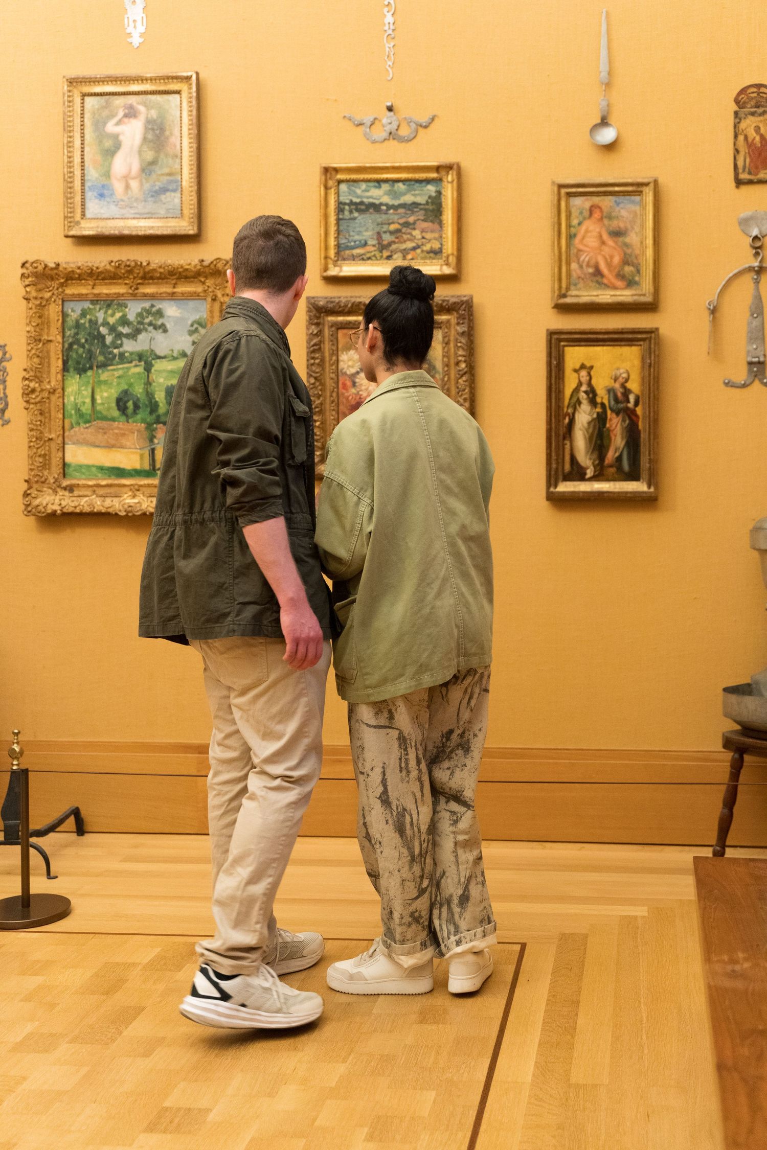 Two visitors in green jackets viewing framed paintings in art museum gallery.