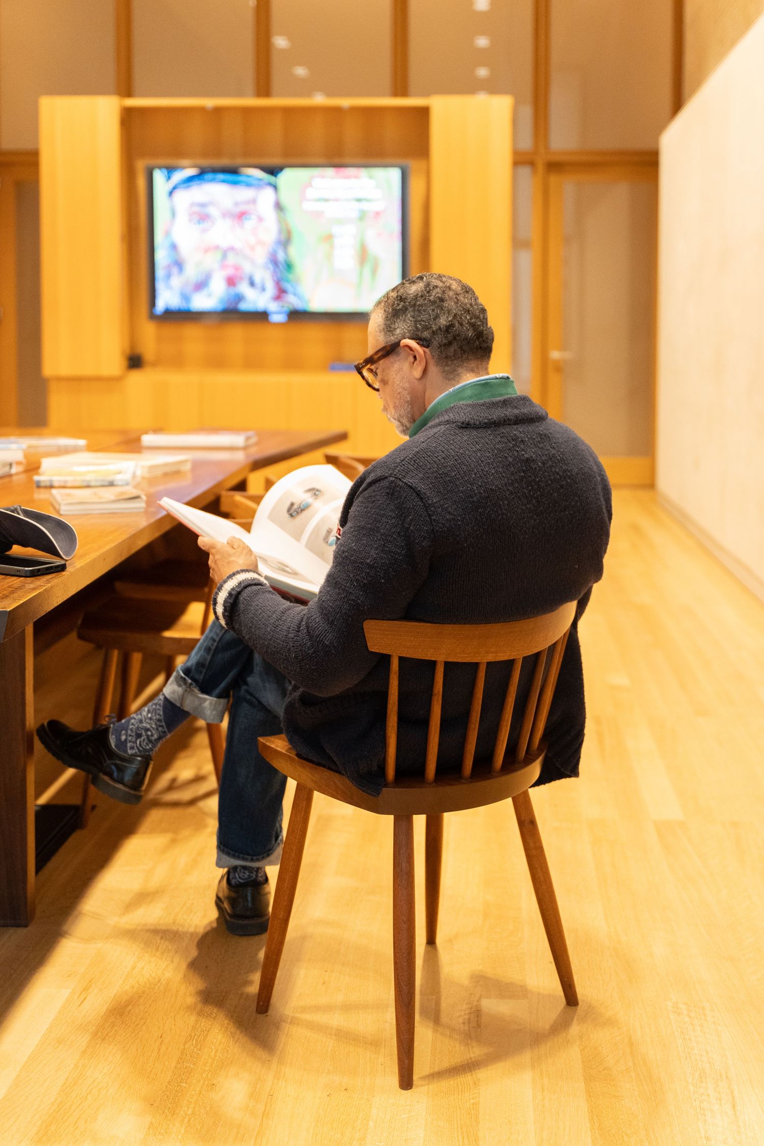 Man in black jacket sits in wooden chair reading document in modern gallery space with TV display.