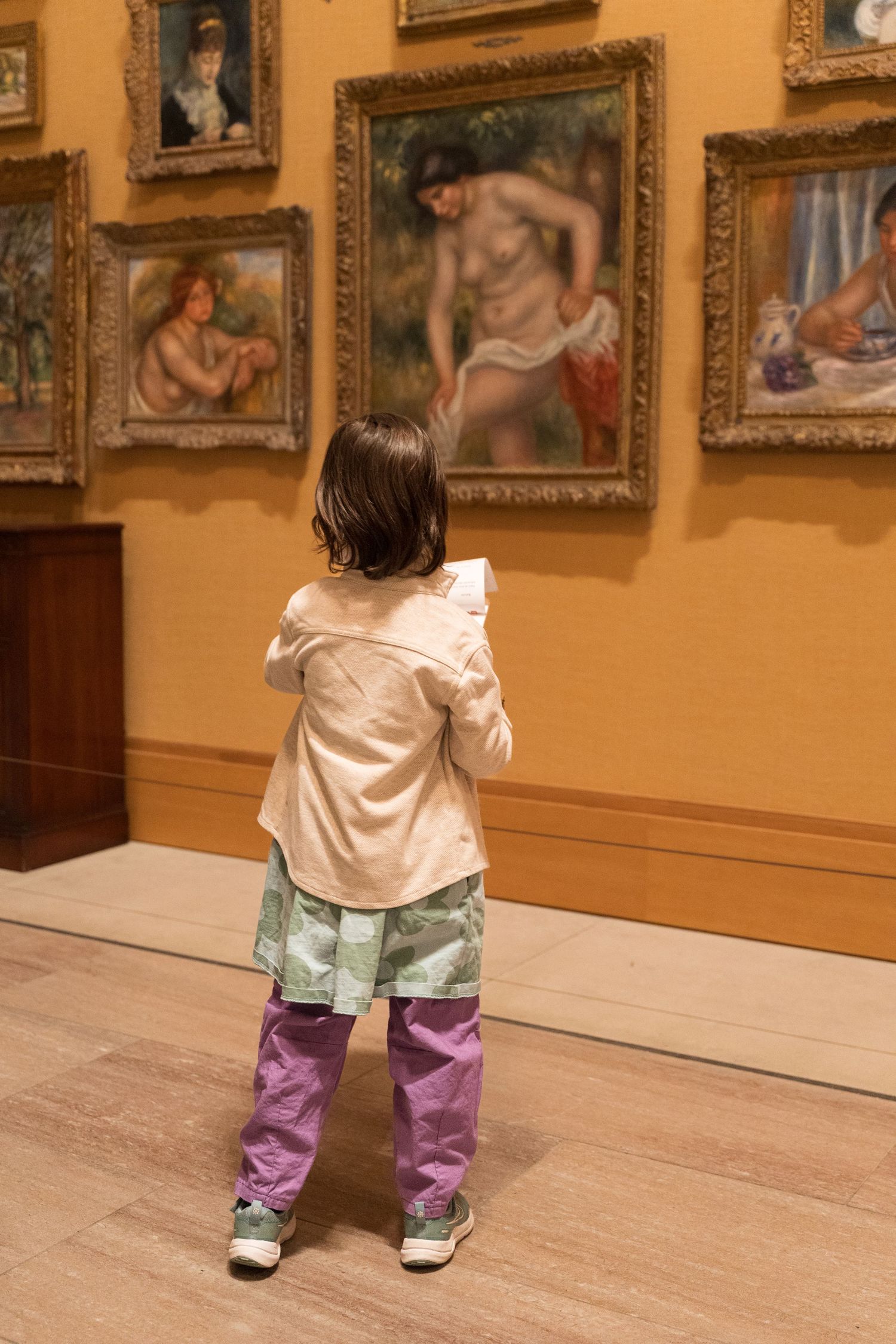 Young child standing alone viewing Renoir paintings in a museum gallery.