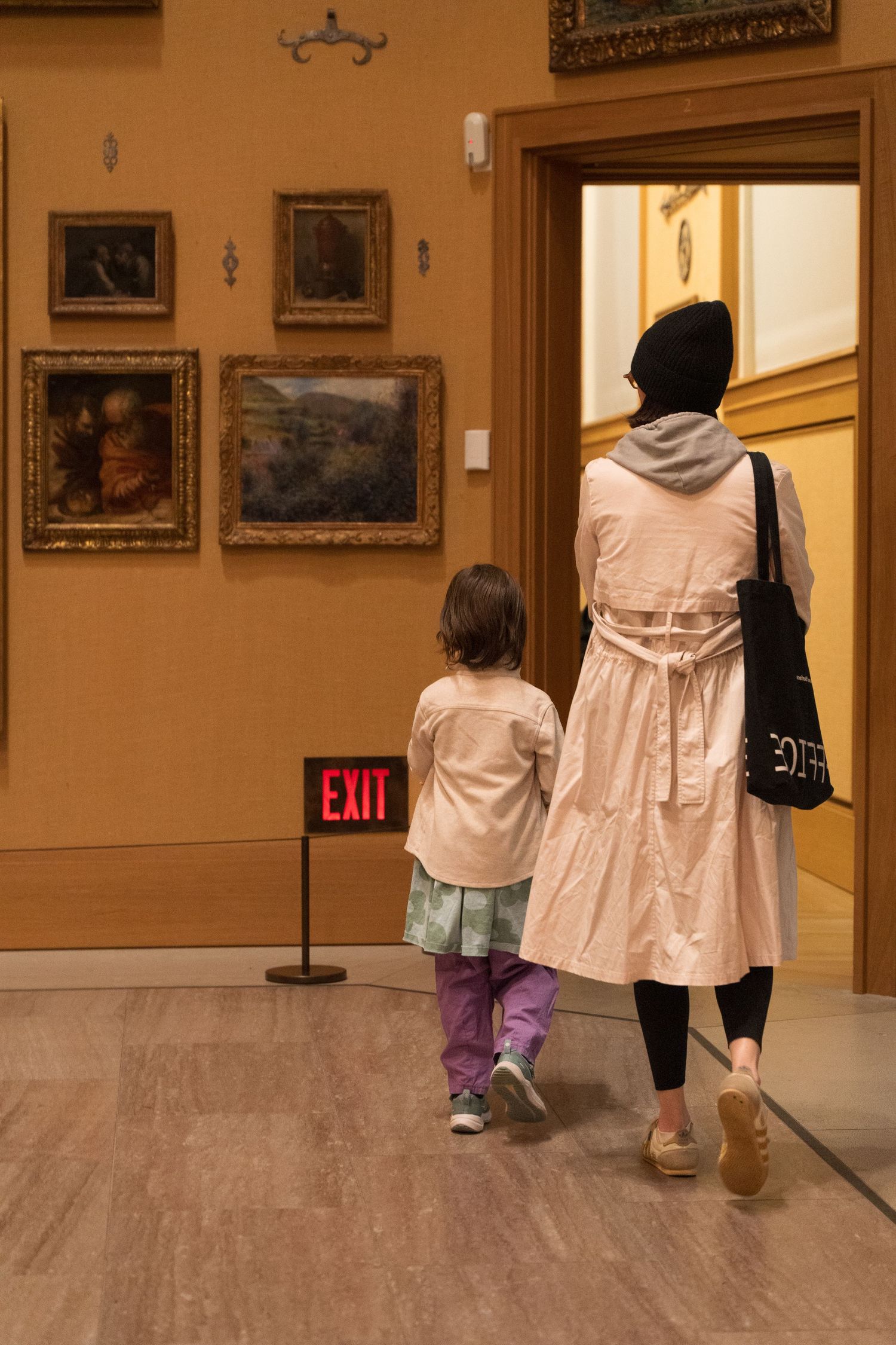 Mother and young child walking hand in hand past paintings in an art museum gallery near an Exit sign.