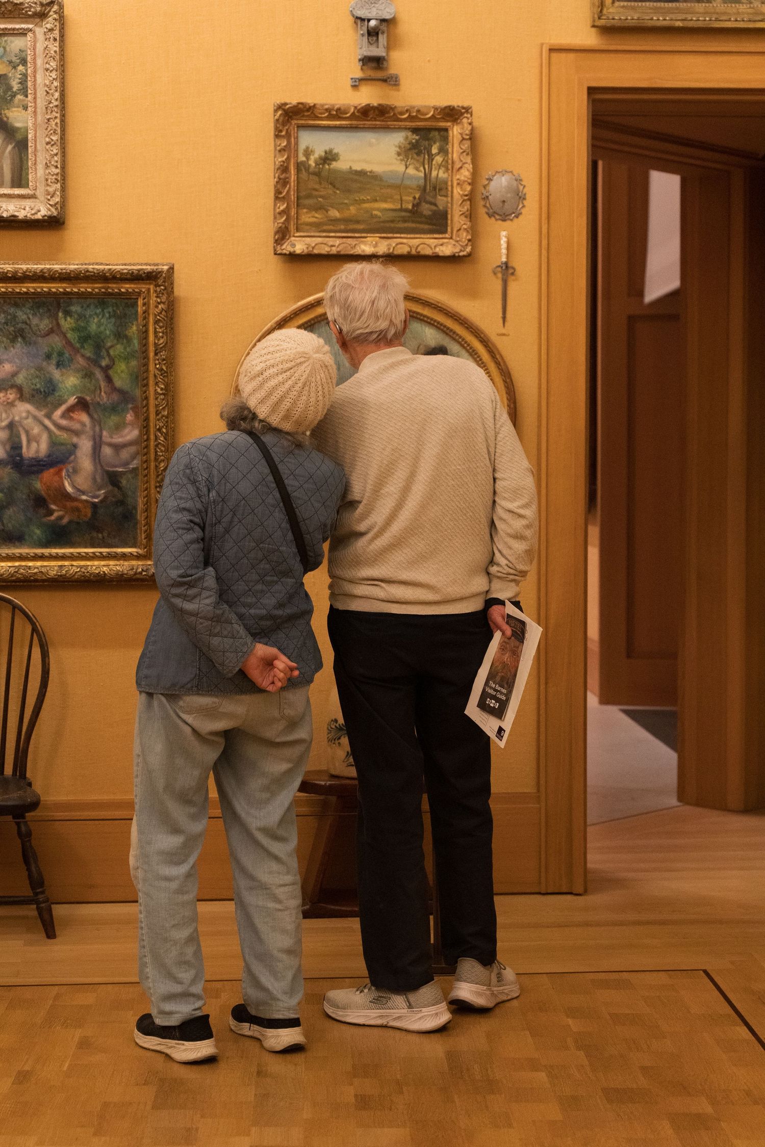 Elderly couple standing close together viewing paintings in a warmly lit art museum gallery.