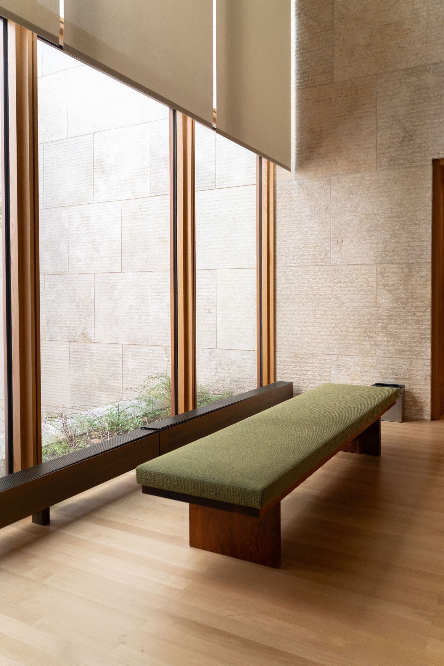 Modern museum interior with a green upholstered bench, wood-framed windows, and natural stone walls.