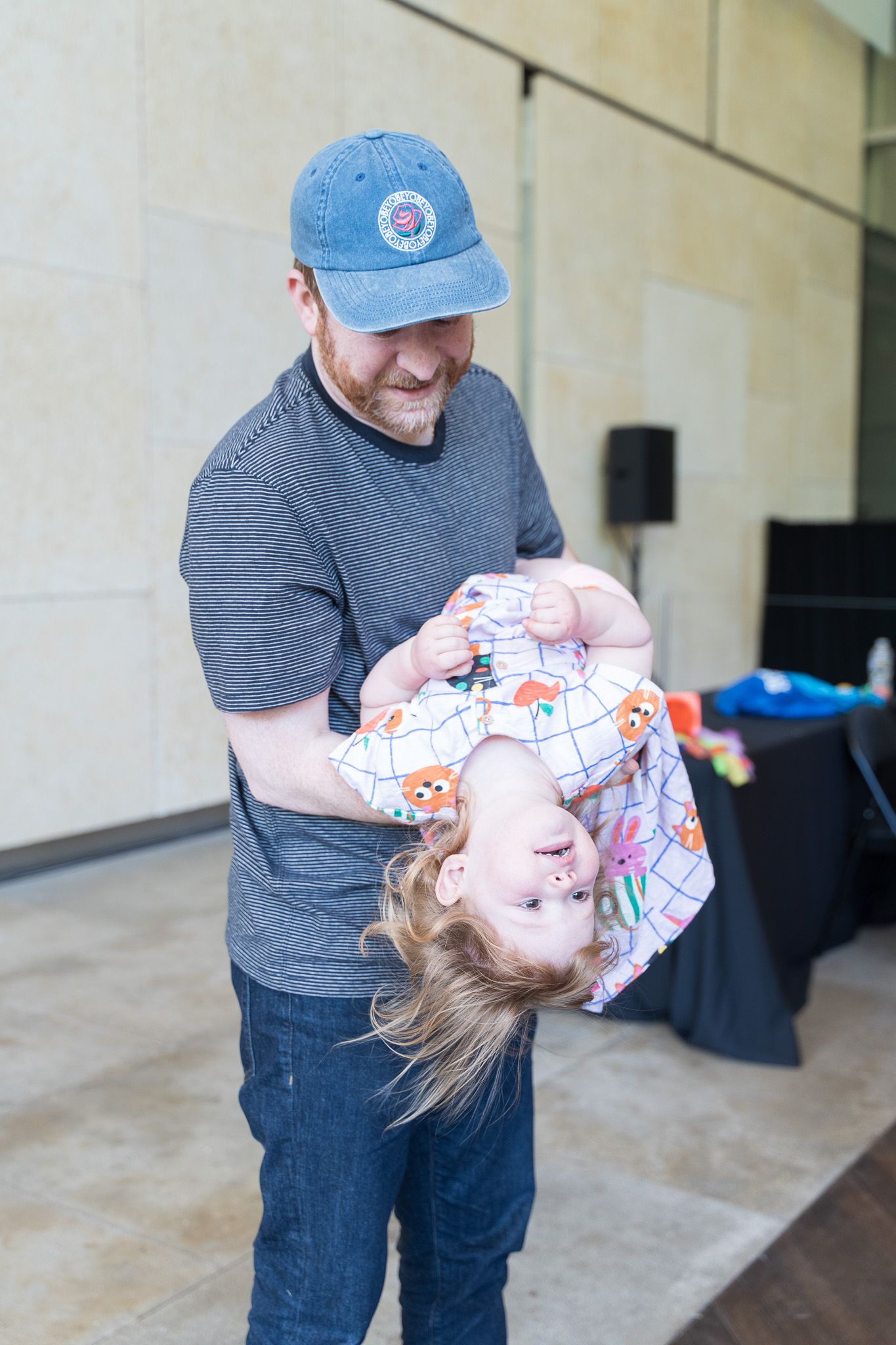 A smiling man in a gray t-shirt and blue cap playfully holds a laughing toddler upside down indoors.