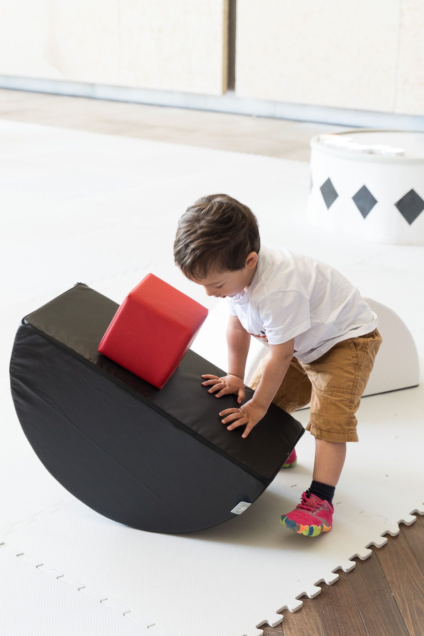 Toddler boy playing with a black foam rocker balance toy with a red block on top in a bright indoor play area.