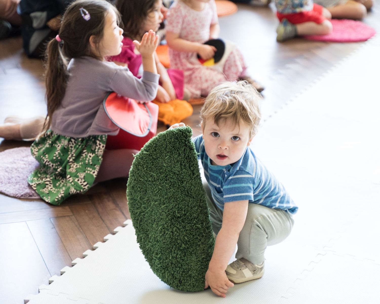 Young children sit on foam mats during a classroom activity while a toddler boy crouches beside a green felt leaf prop.