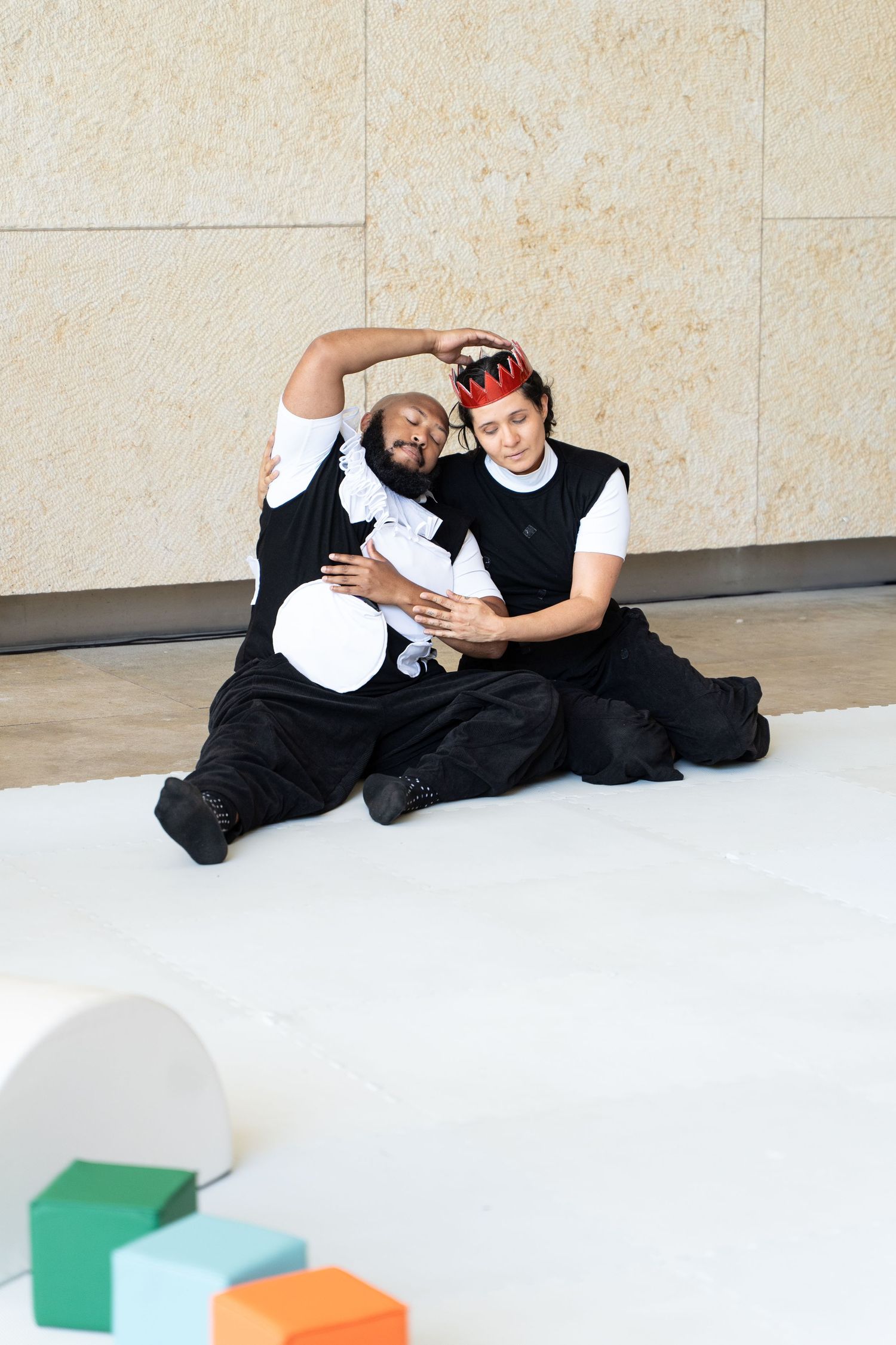 Two performers in black costumes sit on white floor, one wearing a red crown, posing dramatically near colorful blocks.