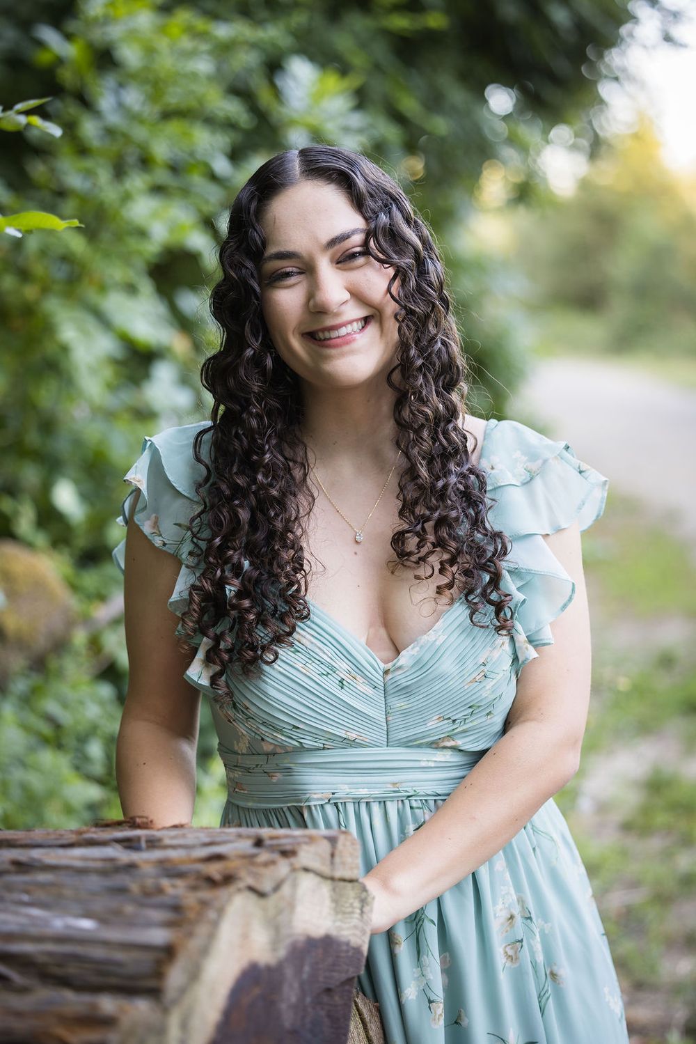 Young woman with curly hair smiling outdoors in a mint floral dress leaning on a log.
