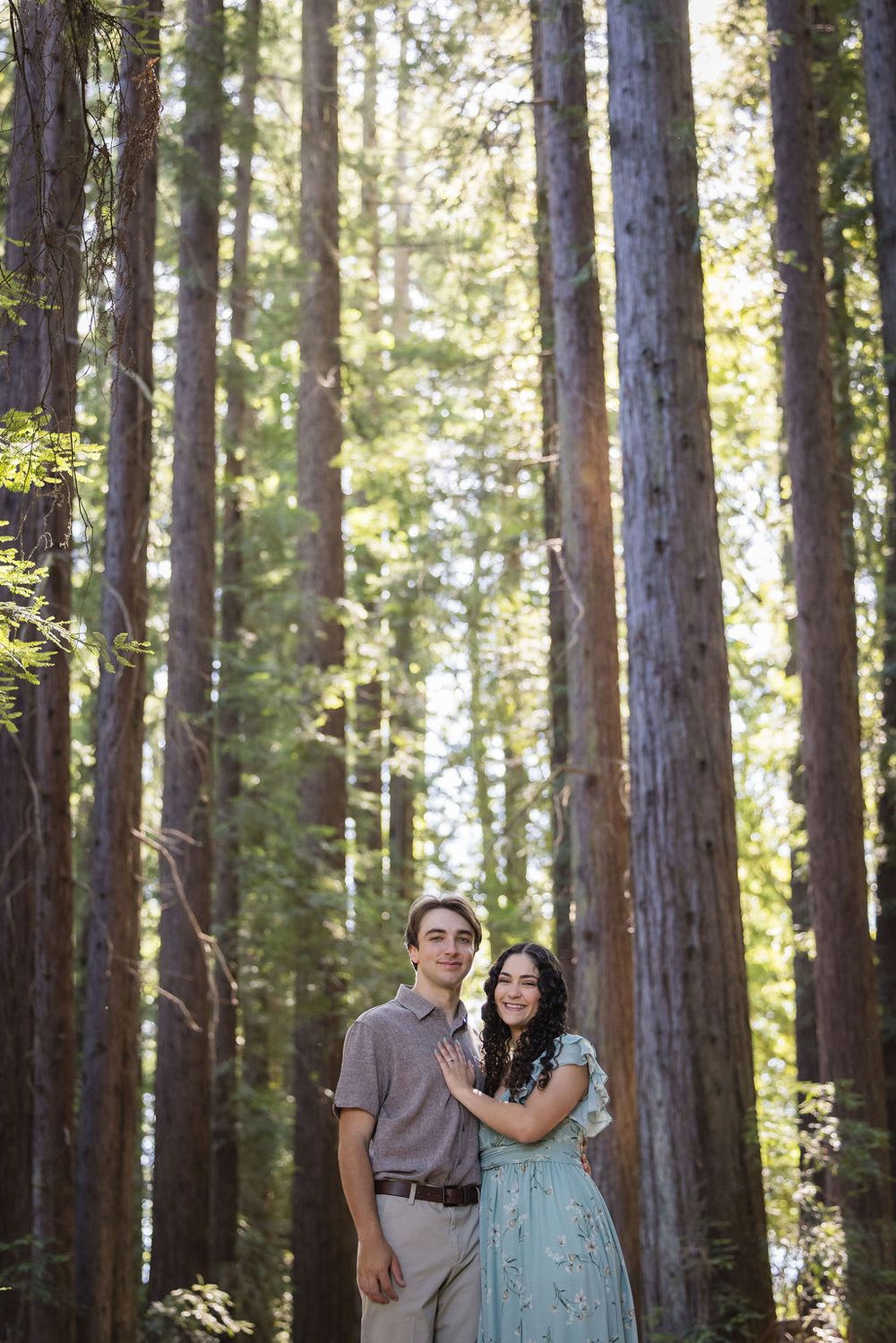 Couple posing among tall redwood trees in a forest, woman in teal dress and man in gray shirt.