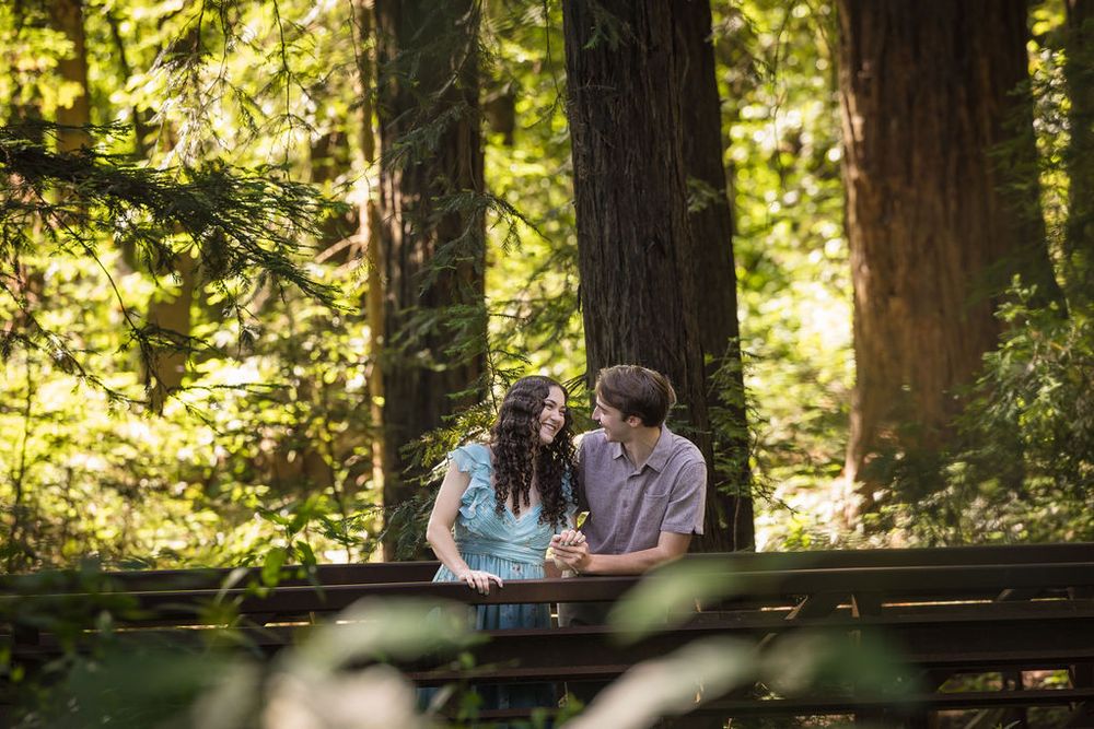 Couple sharing a romantic moment on a wooden bridge surrounded by towering redwood trees in a lush forest.