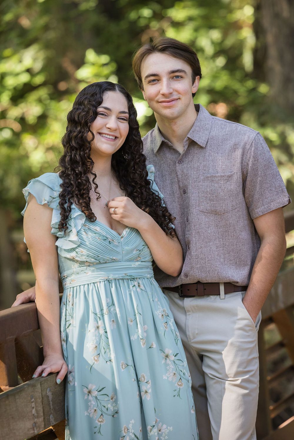 Young couple posing together outdoors on a wooden bridge, woman in floral mint dress and man in gray shirt.