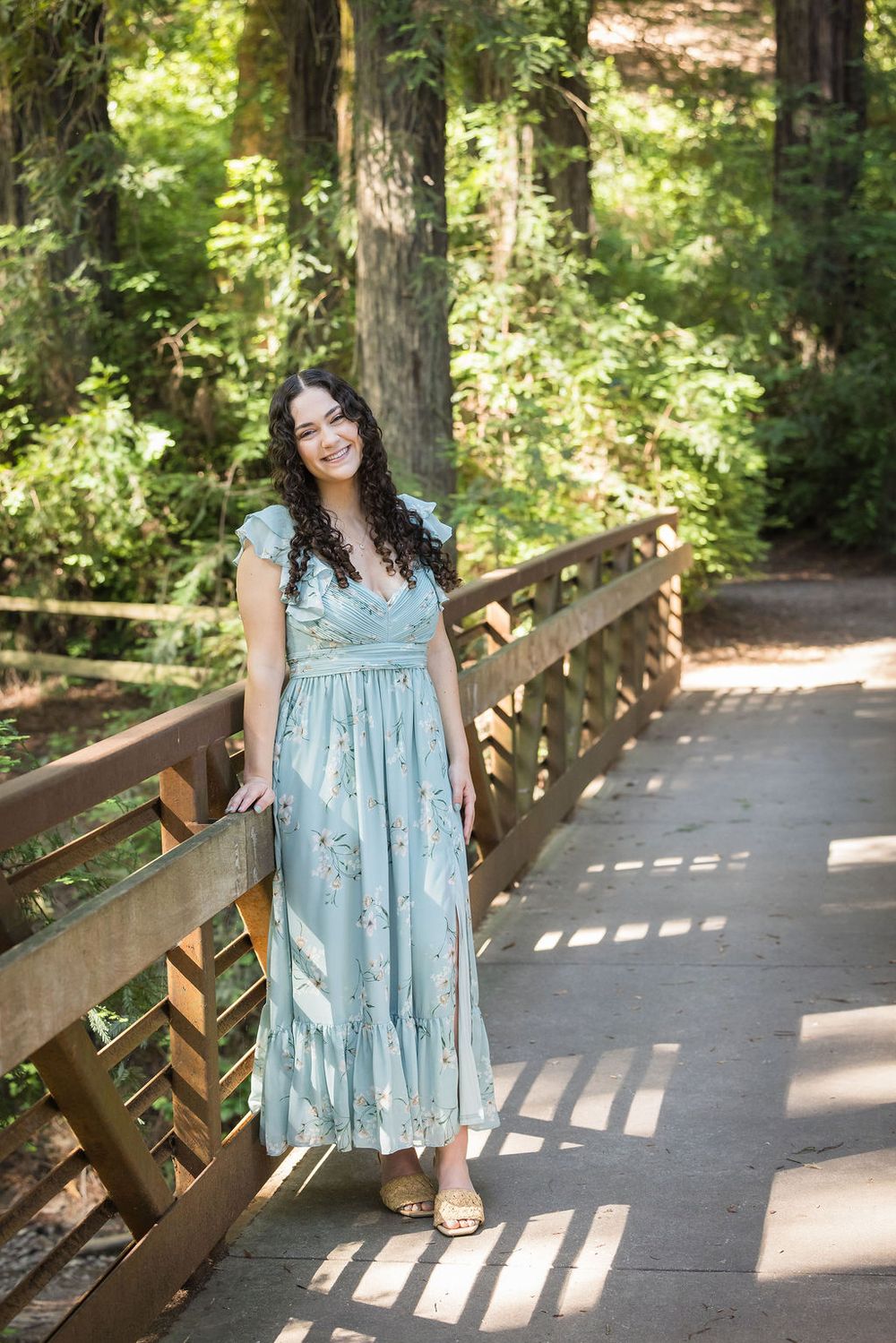 Woman in light blue floral maxi dress poses on wooden bridge surrounded by lush green forest.