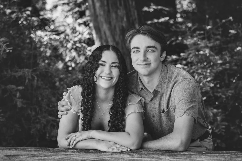 Black and white portrait of a smiling young couple leaning on a wooden surface outdoors among trees.
