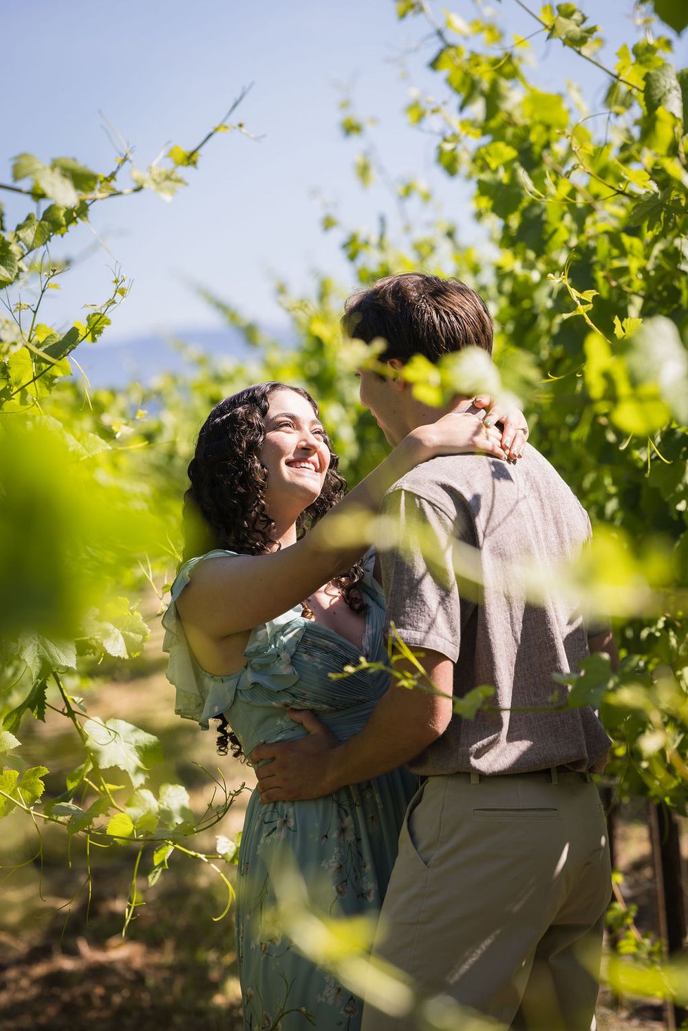 Happy couple embracing and smiling among green grapevines in a sunny vineyard on a clear day.