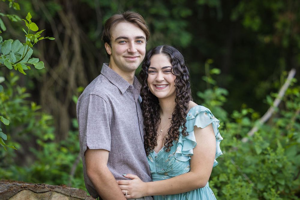 Smiling couple poses together outdoors, woman in teal ruffled dress and man in gray shirt.