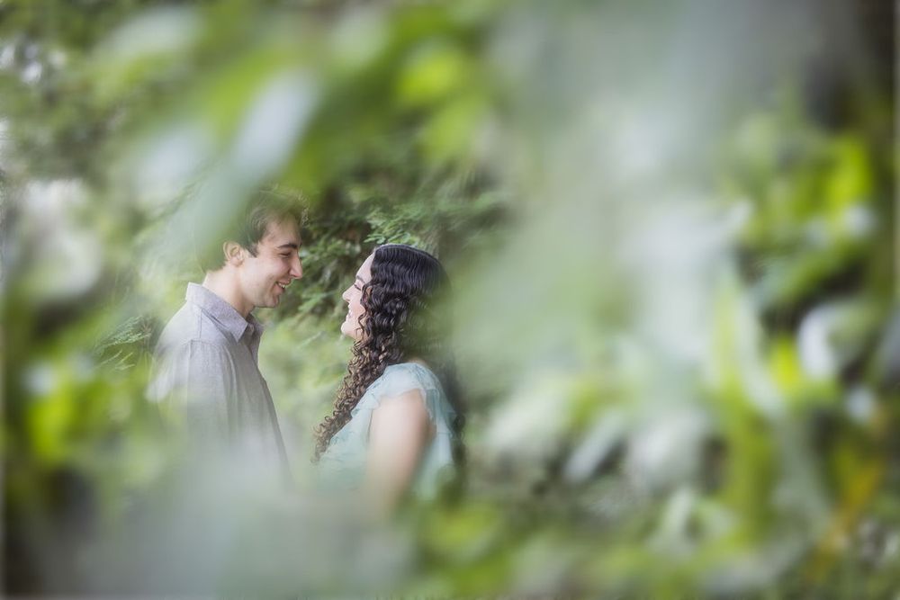 Romantic couple gazing at each other framed by soft green foliage in a dreamy outdoor setting.