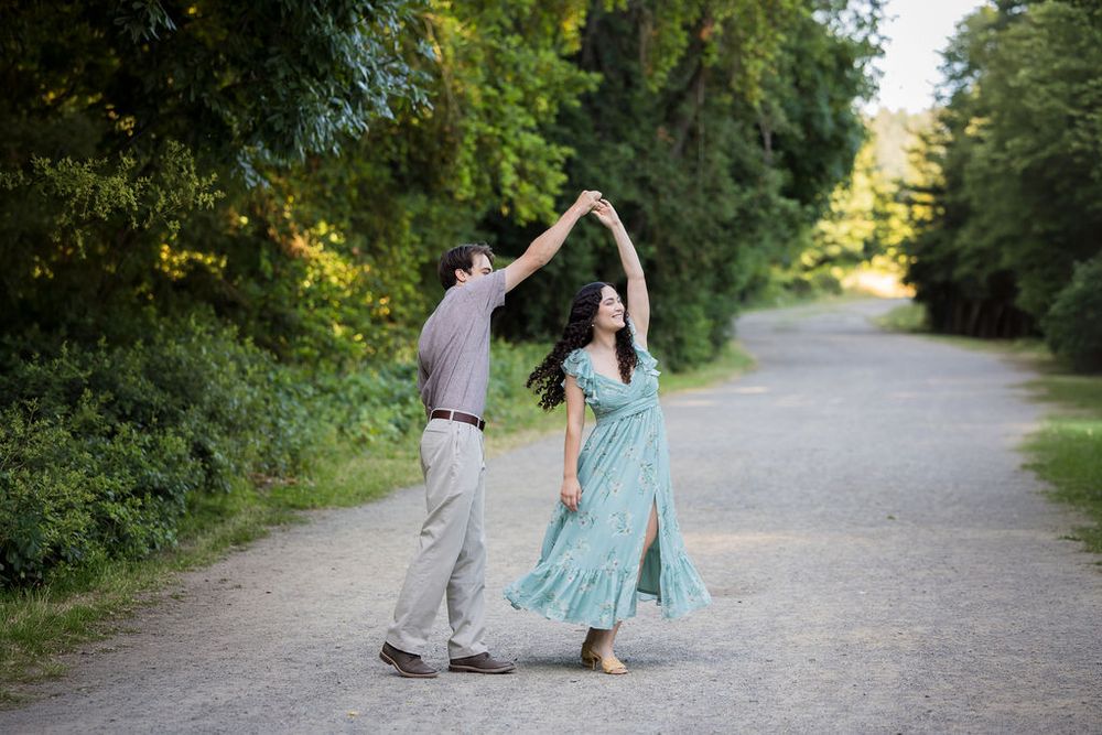Couple dancing on a tree-lined path, woman in flowing teal dress being twirled by her partner.