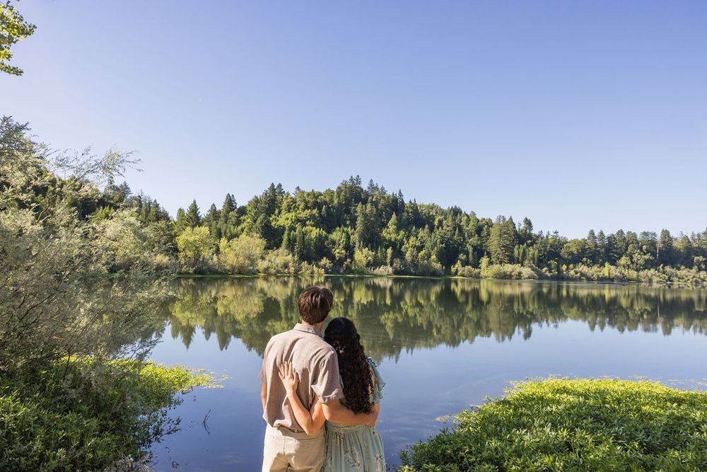 A couple stands arm in arm gazing at a serene lake reflecting green forested hills under clear blue sky.