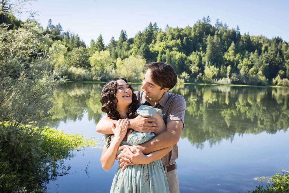 Happy couple embracing and laughing by a calm reflective lake surrounded by green forested hills.