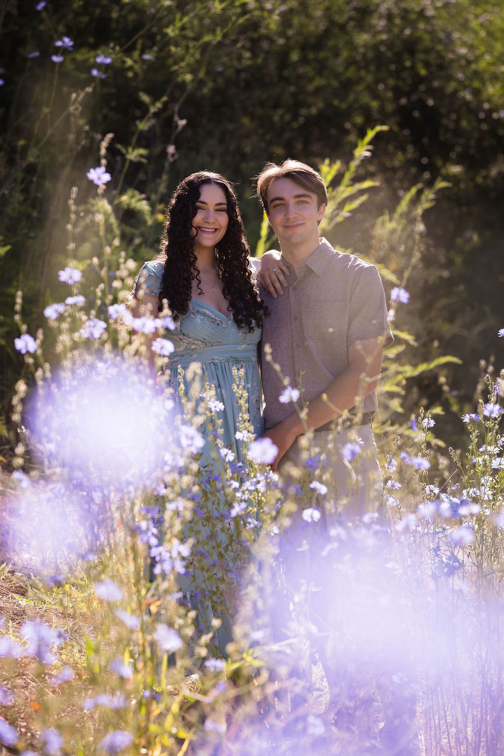 Smiling couple standing together in a sunlit field of purple wildflowers.