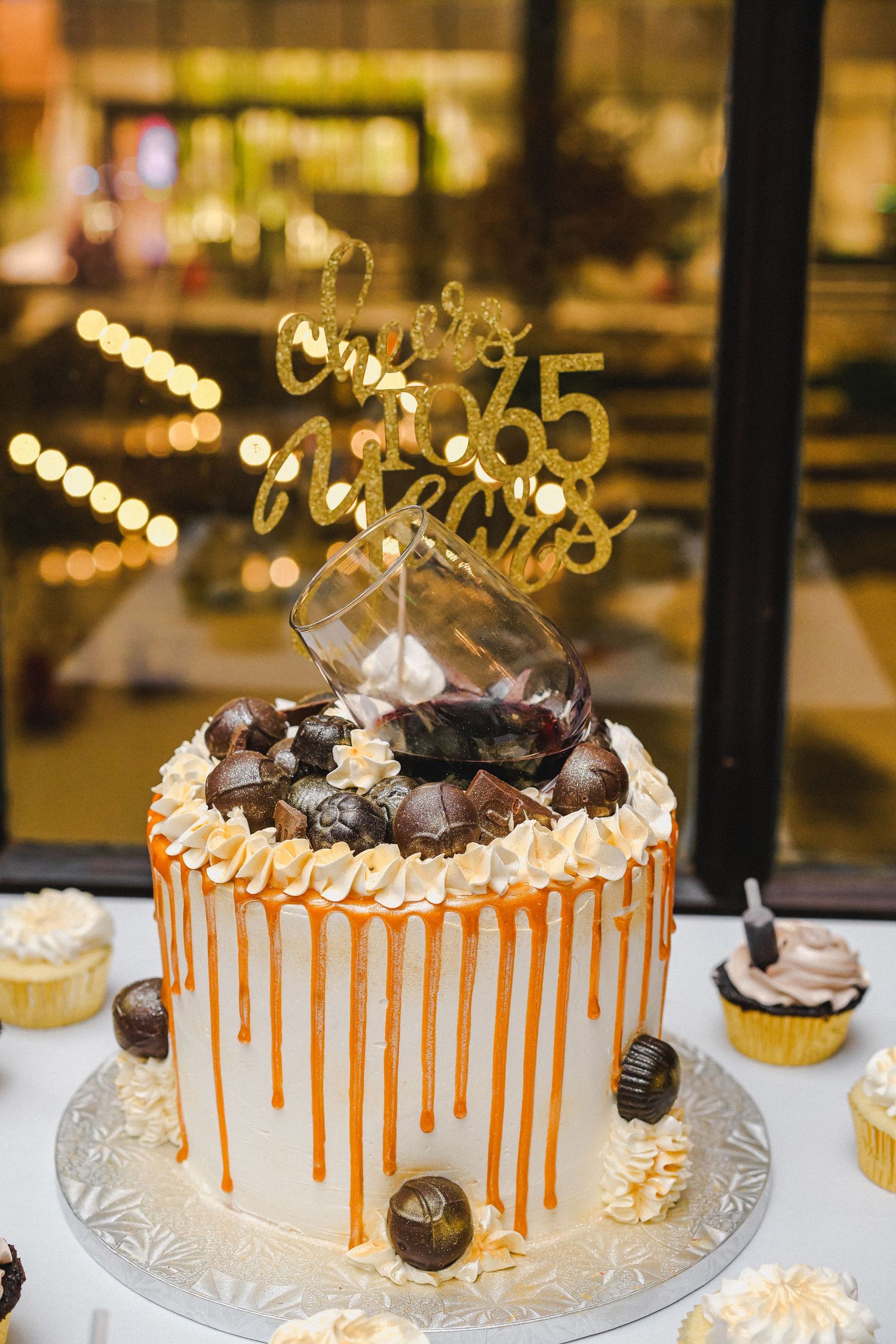 Elegant 65th birthday cake with caramel drip, chocolate truffles, whipped cream, gold topper, and a decorative tipped glass on top.