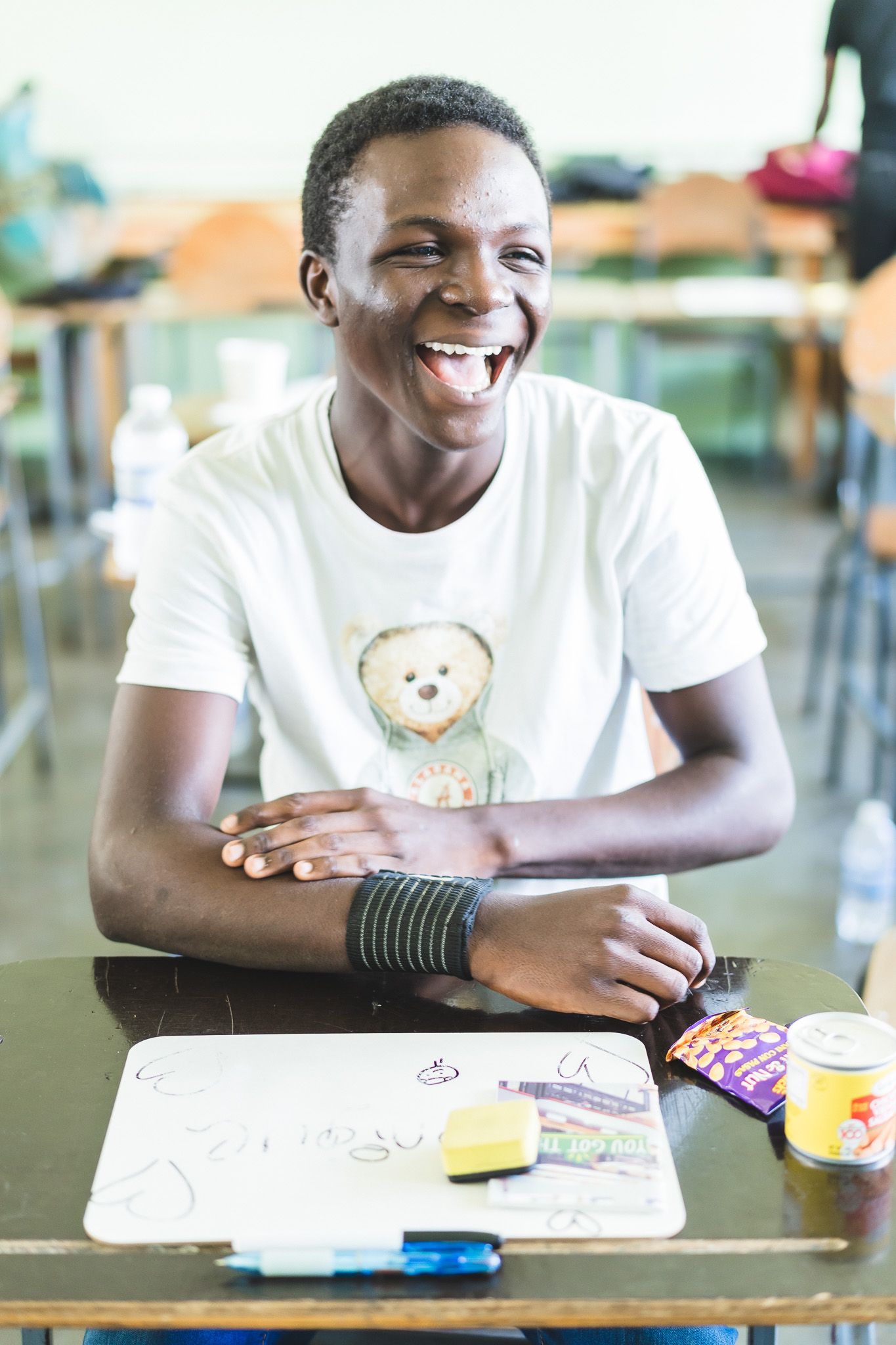 Laughing young man in white bear t-shirt sits at desk with whiteboard and markers in bright classroom.