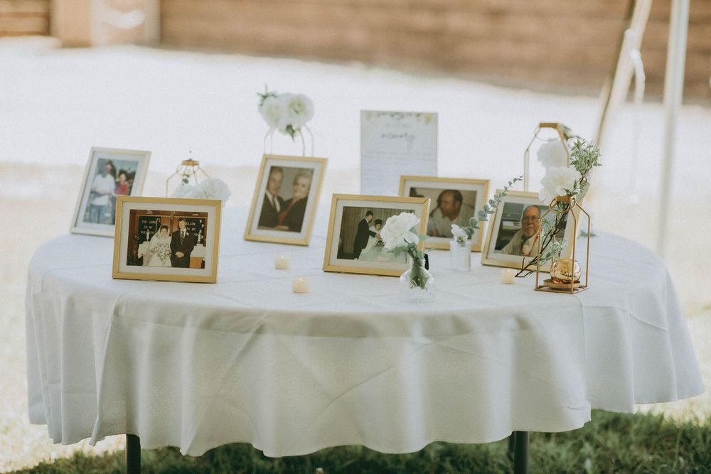 Elegant wedding memory table with gold-framed family photos, white flowers, and soft linen tablecloth displayed outdoors.