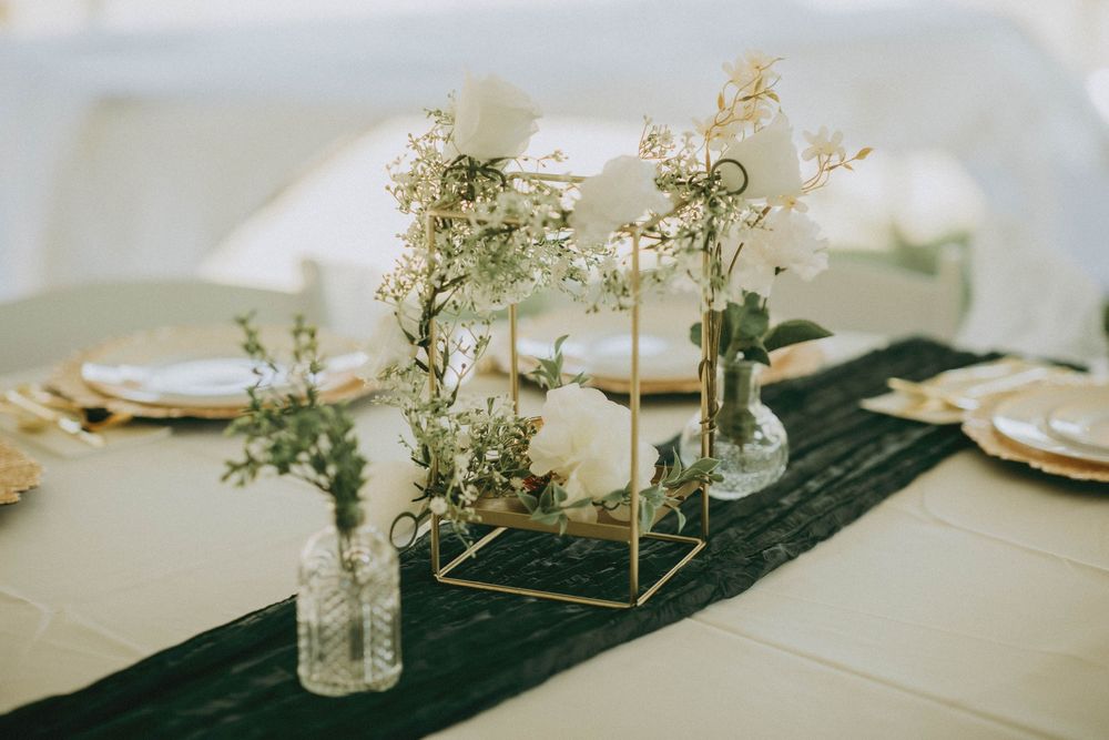 Elegant wedding table centerpiece with gold geometric frame, white flowers, and greenery on a dark runner.