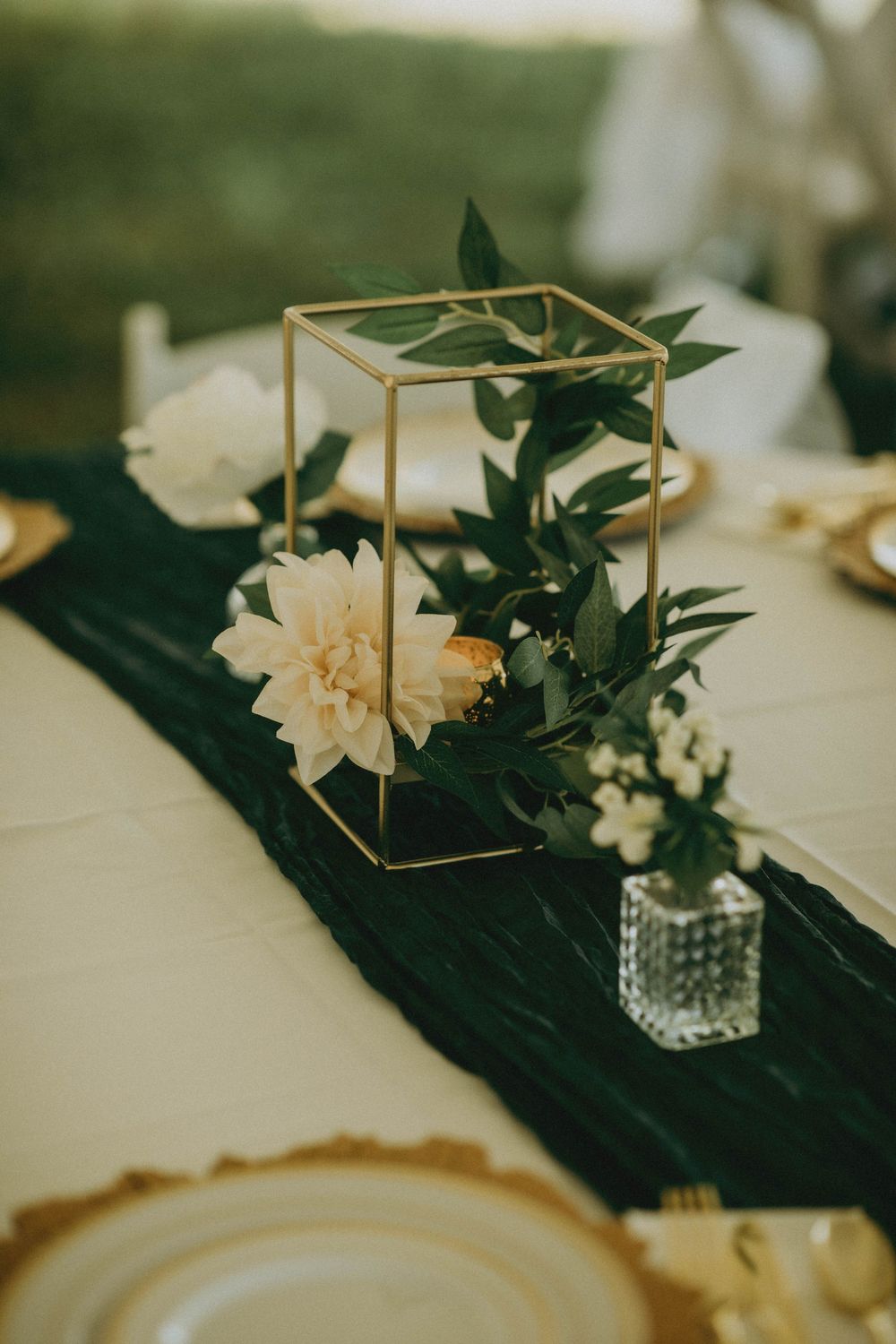 Gold geometric centerpiece with white dahlia, greenery, and dark runner on elegant table setting.
