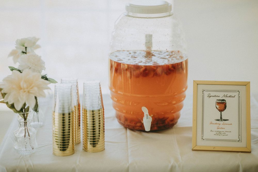Glass beverage dispenser filled with orange punch on a white tablecloth beside gold cups and white flowers.