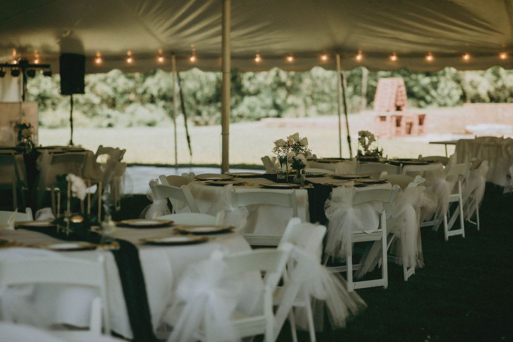 Elegant outdoor wedding reception under a tent with white chairs, draped tables, string lights, and floral centerpieces.