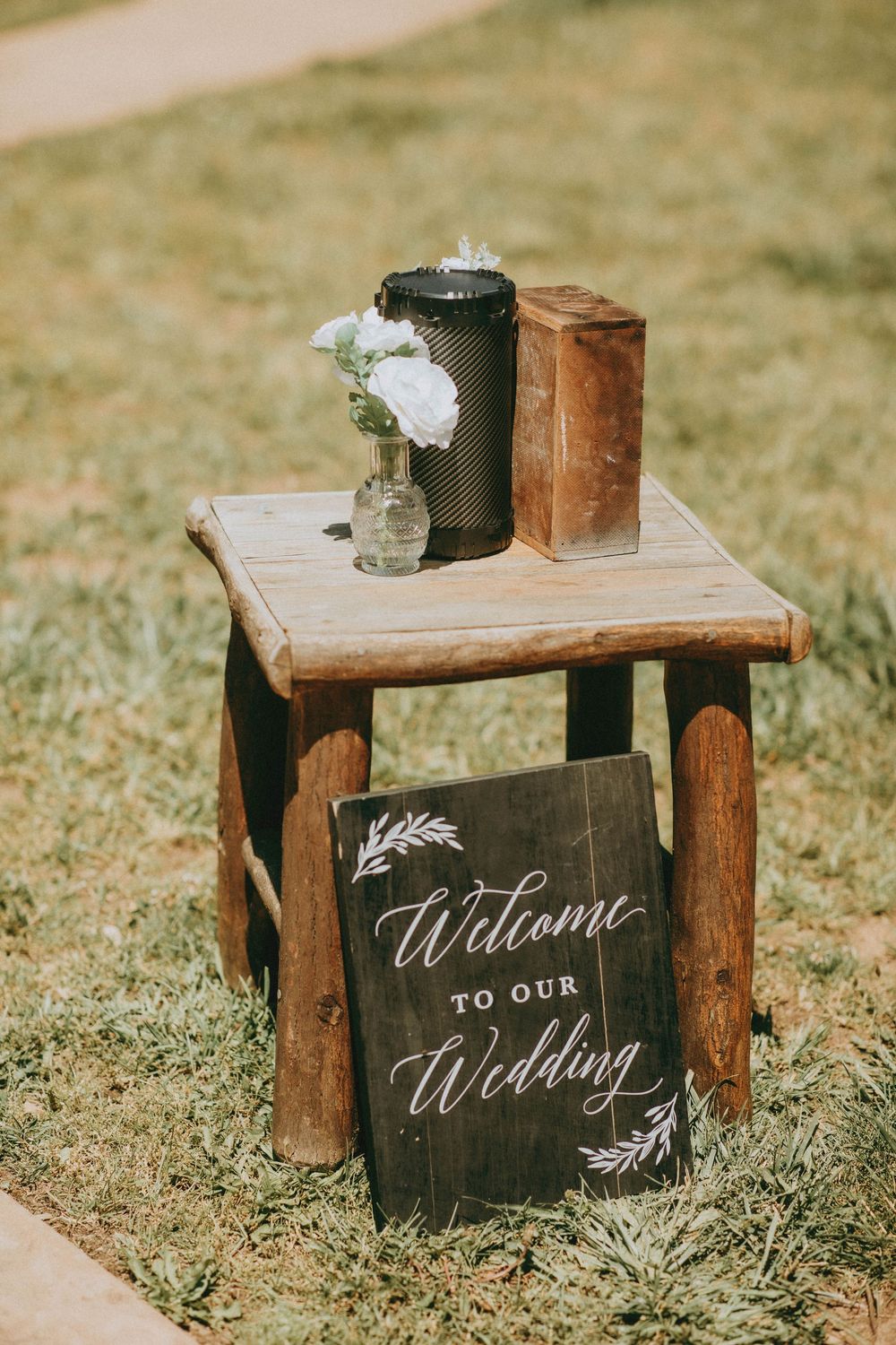 Rustic wooden stool with a Welcome to Our Wedding chalkboard sign, small floral vase, and vintage decor on green grass.