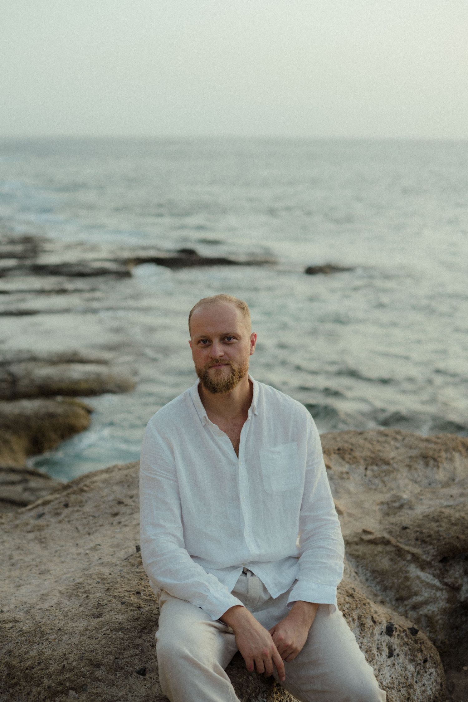 Bald man with beard in white linen shirt sitting on rocky coastal shore with calm ocean in background.