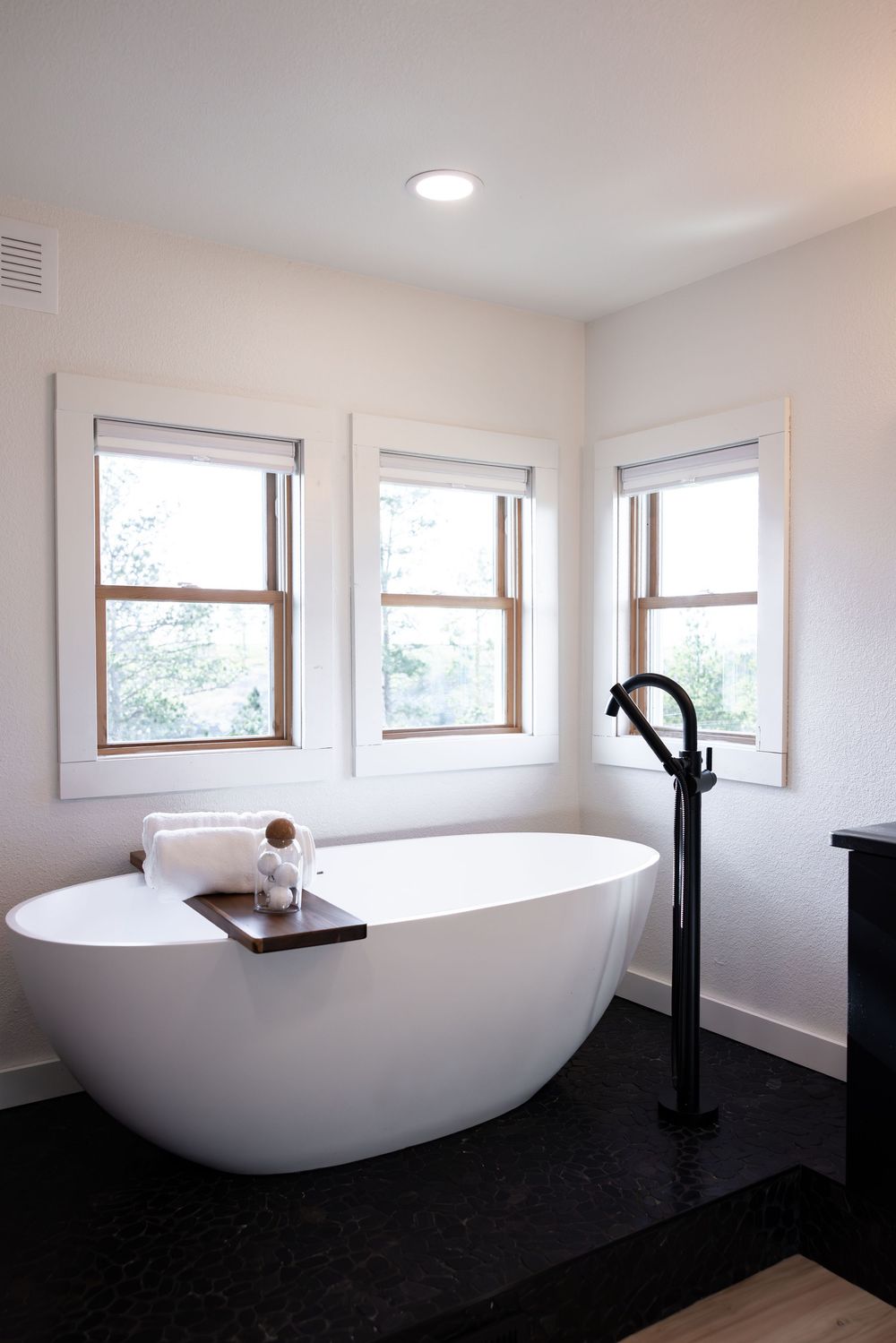 Elegant freestanding white soaking tub with matte black floor faucet on dark tile, flanked by three bright windows.