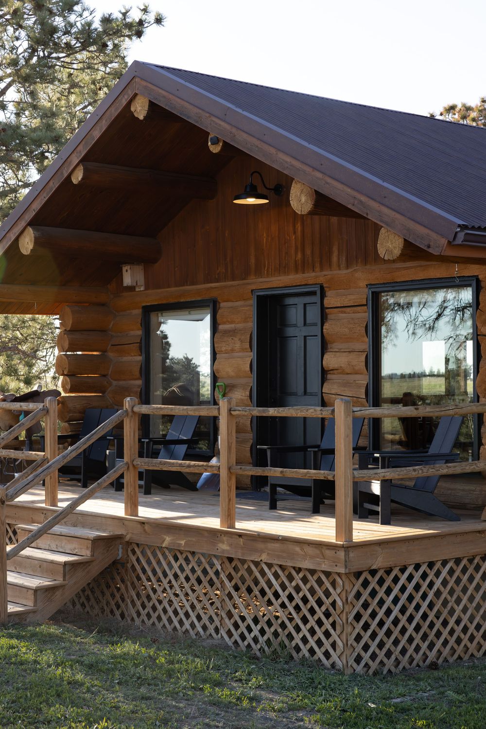 Rustic log cabin with wooden deck, Adirondack chairs, large windows, and brown metal roof surrounded by trees.