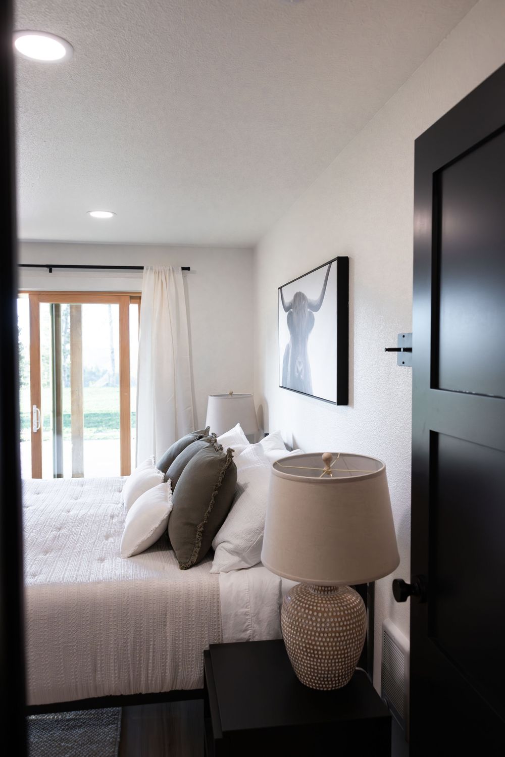 Modern bedroom with white bedding, gray pillows, bedside lamp, framed wall art, and sliding glass doors with white curtains.