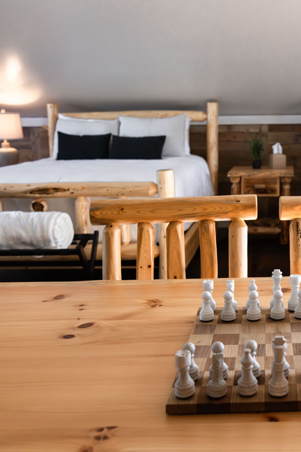 Rustic log cabin bedroom with chess set on wood table, natural timber bed frame, and cozy white bedding.