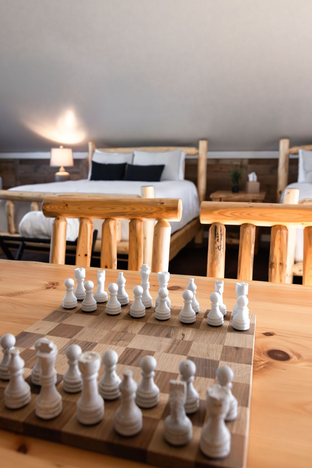 A chess board with white pieces set up on a wooden table in a rustic log cabin bedroom with natural wood furniture.