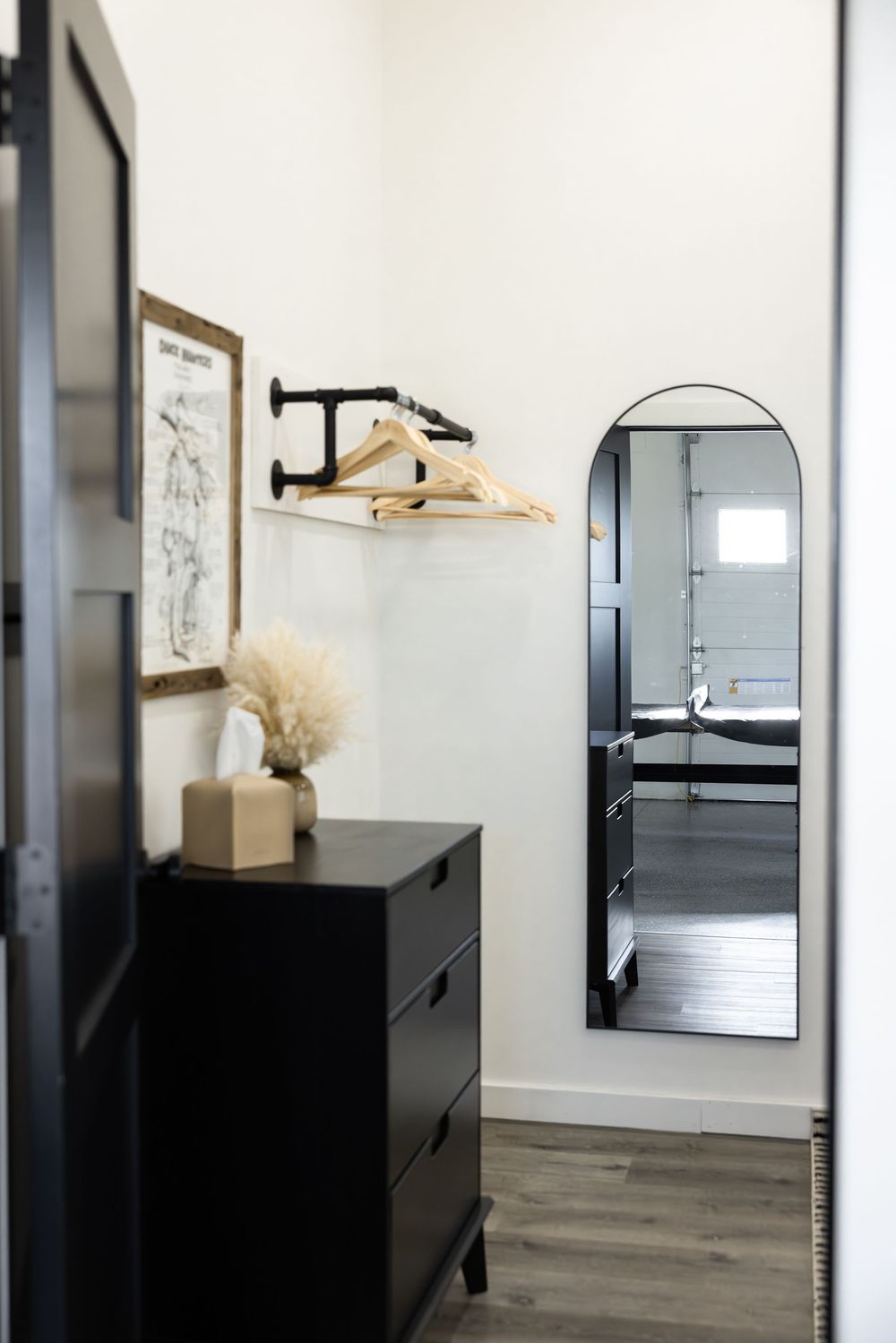Modern entryway with black dresser, arched mirror, wooden hangers on wall shelf, and framed art in minimalist black and white decor.