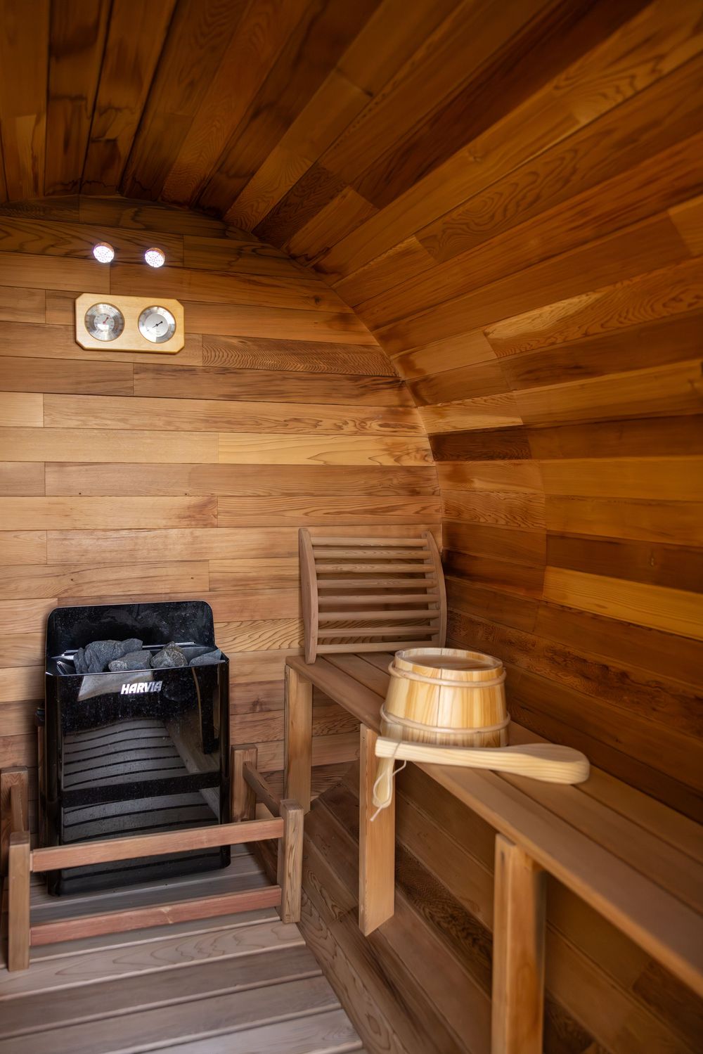 Interior of a cedar barrel sauna with wooden benches, Harvia heater, bucket, and ladle under curved ceiling lights.