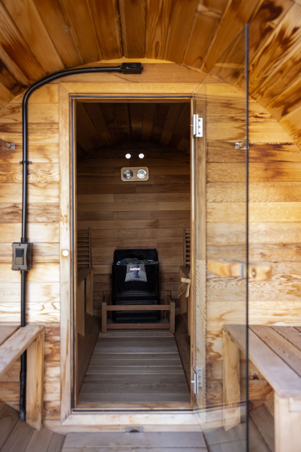Interior view of a wooden barrel sauna with bench seating, glass door, and black sauna heater inside.