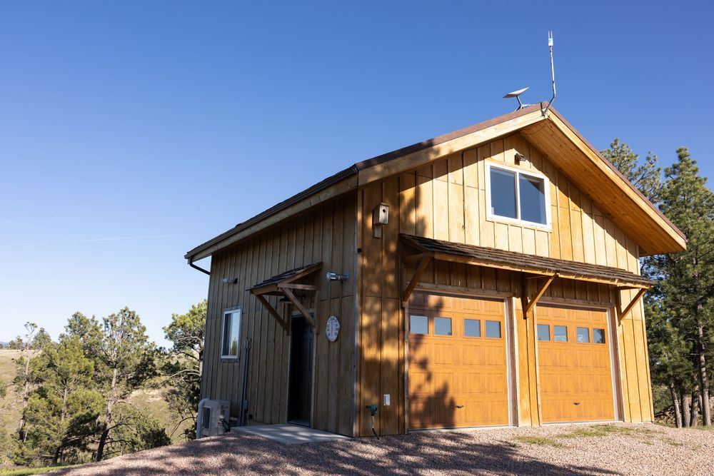 Rustic wooden barn-style building with cedar siding, double garage doors, and a gambrel roof under a clear blue sky.