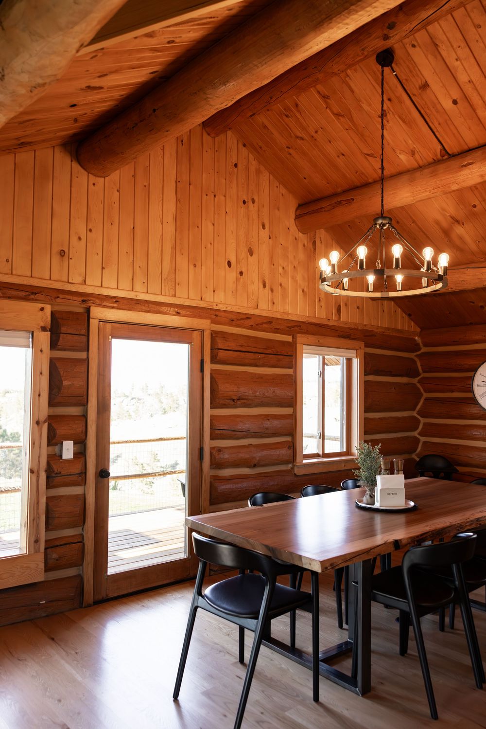 Rustic log cabin dining room with wooden walls, vaulted ceiling, chandelier, and a dark wood table with black chairs.