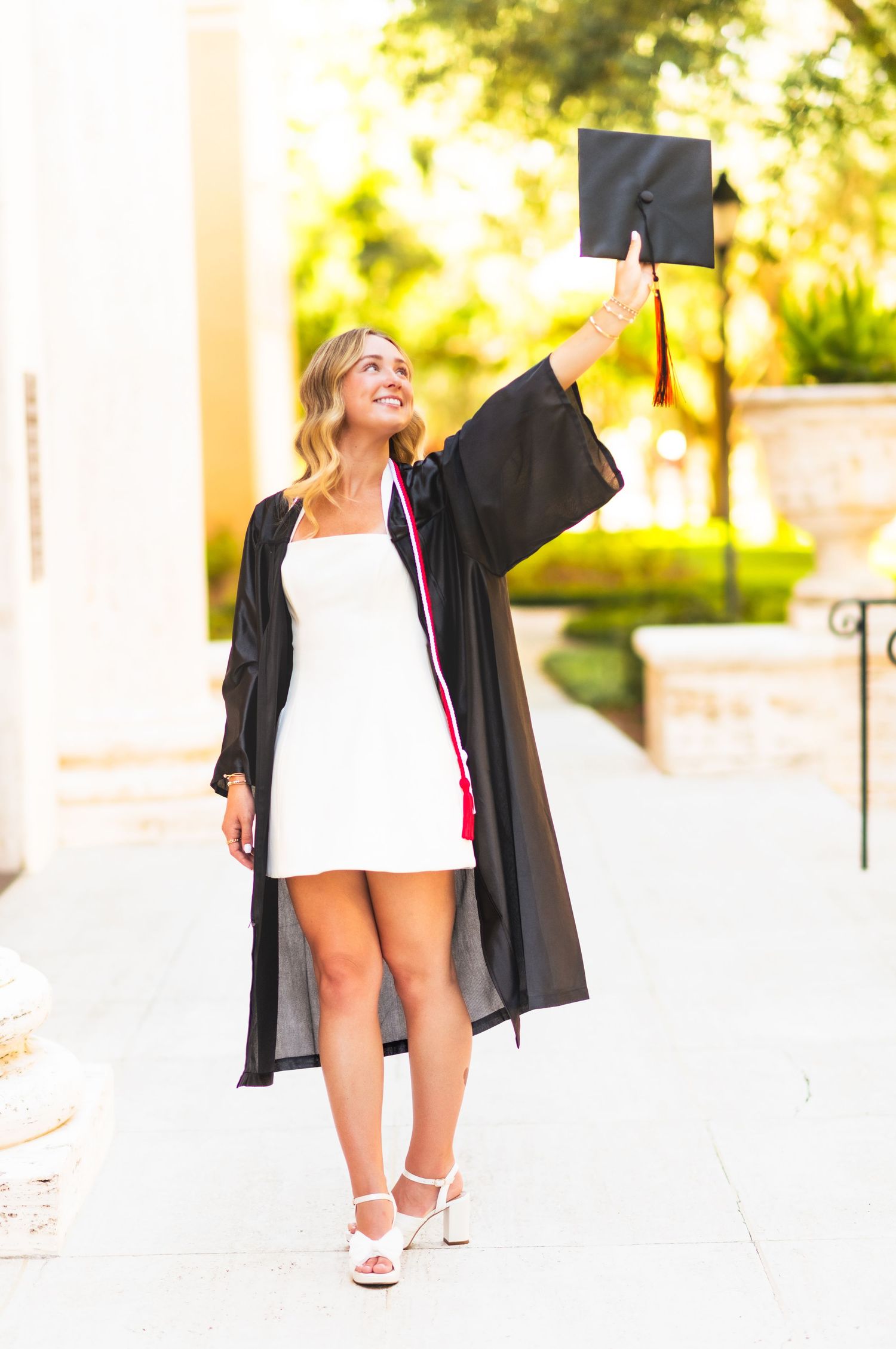 Smiling female graduate in black gown and white dress holds cap up outdoors.