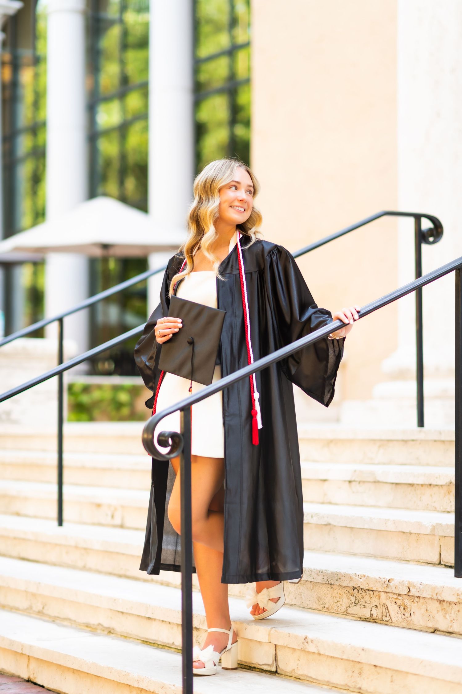 Female graduate in black gown holds cap on sunny campus steps.