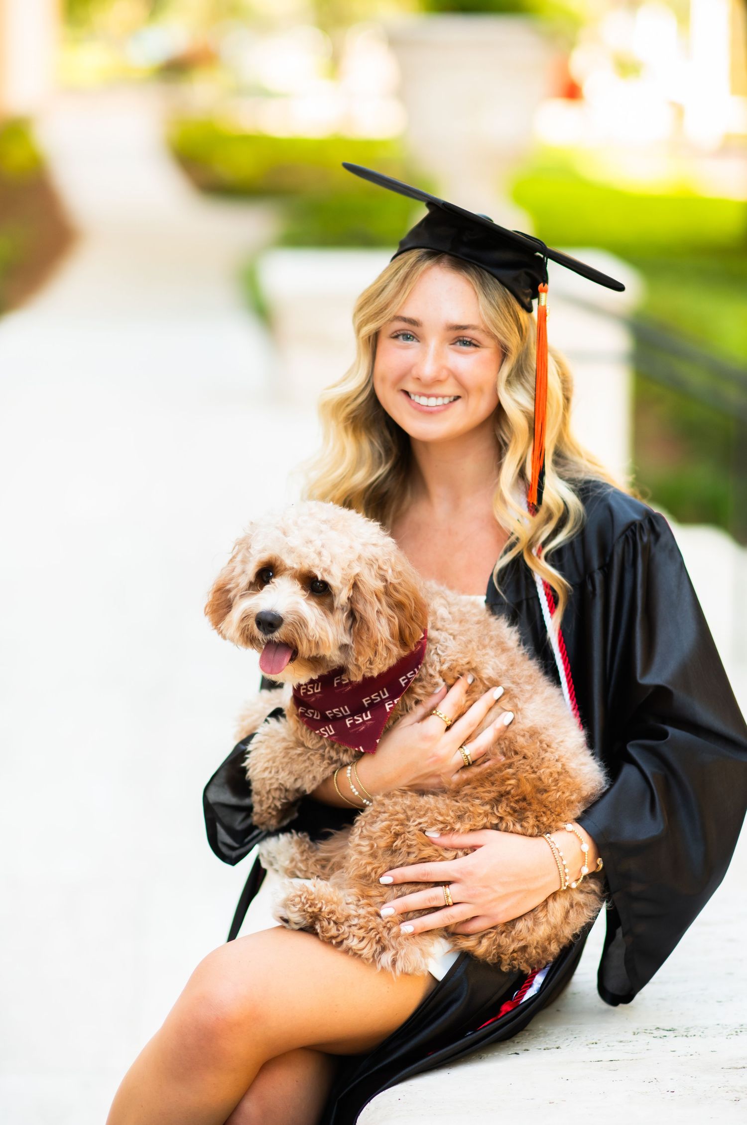 Smiling female graduate in black cap and gown holds fluffy dog outdoors.