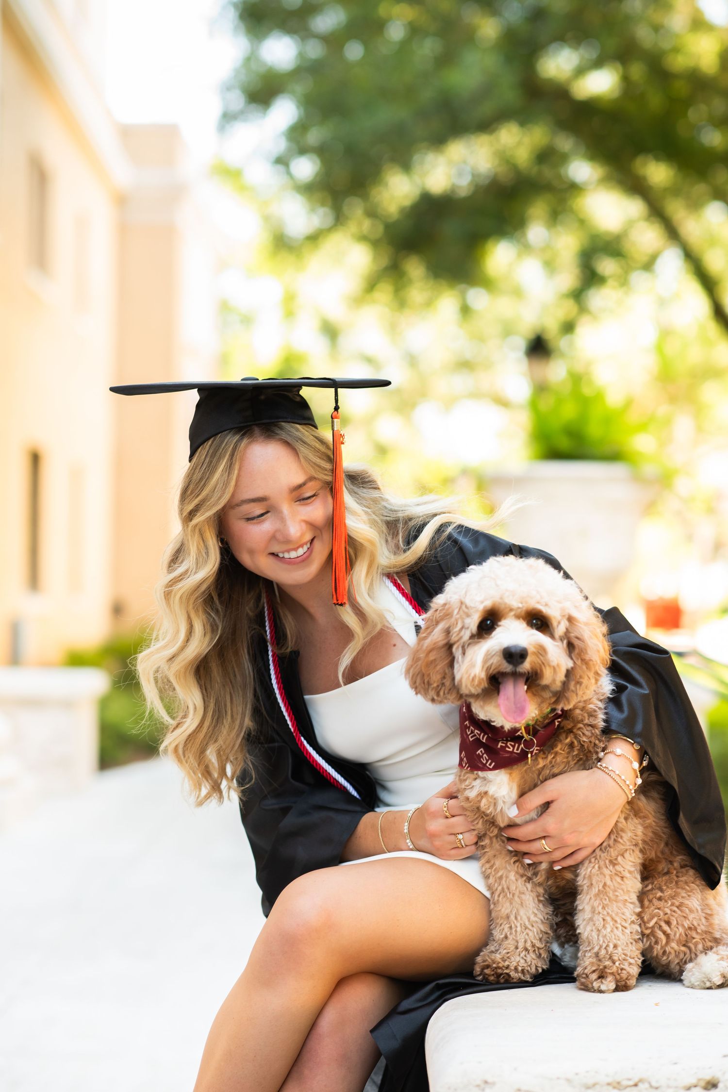 Smiling female graduate in cap and gown holding a fluffy golden doodle dog outdoors on campus.