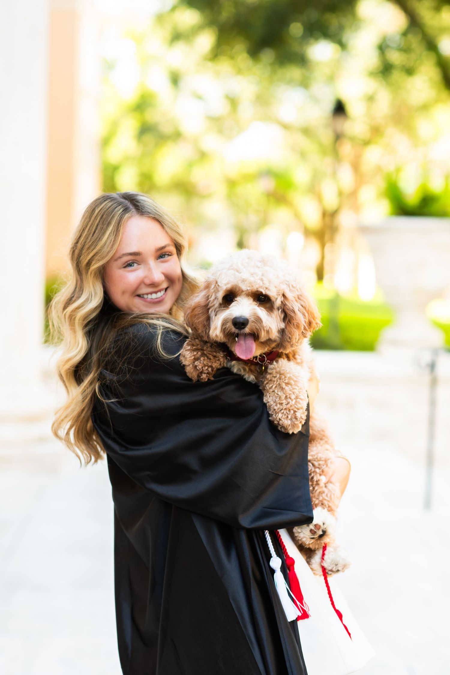 Smiling graduate in black gown holding a fluffy brown dog outdoors.