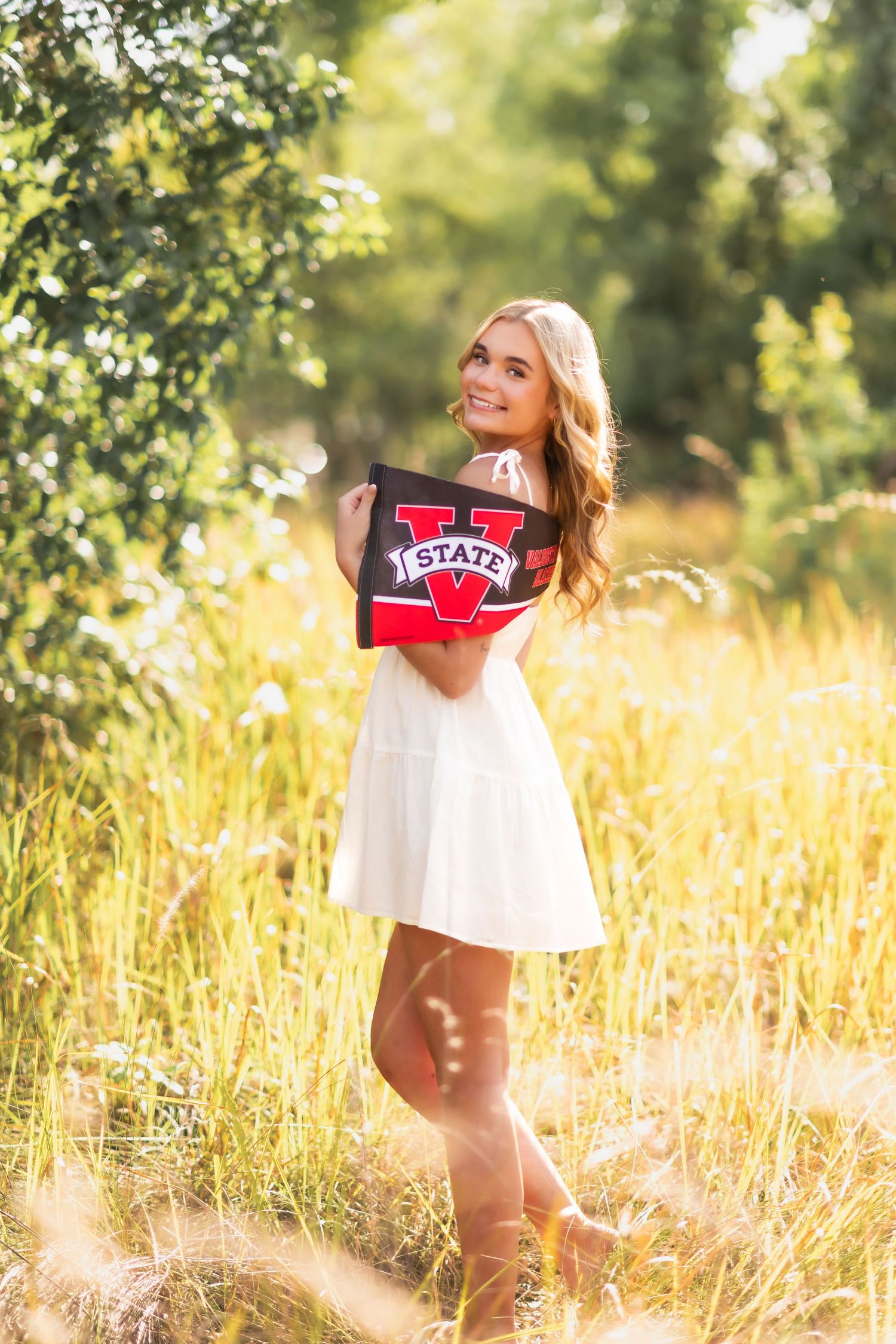 Smiling blonde woman in white dress holding a V State pennant in a sunny golden field.