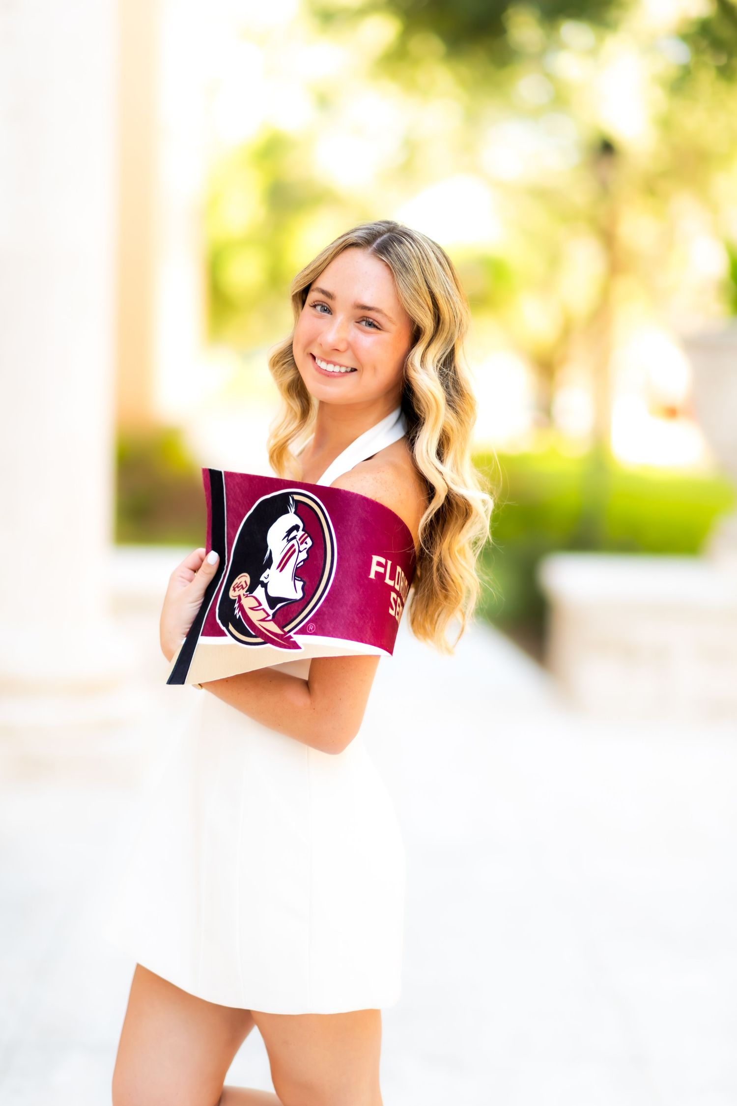 Smiling blonde woman in white dress holding Florida State University flag outdoors on a sunny day.