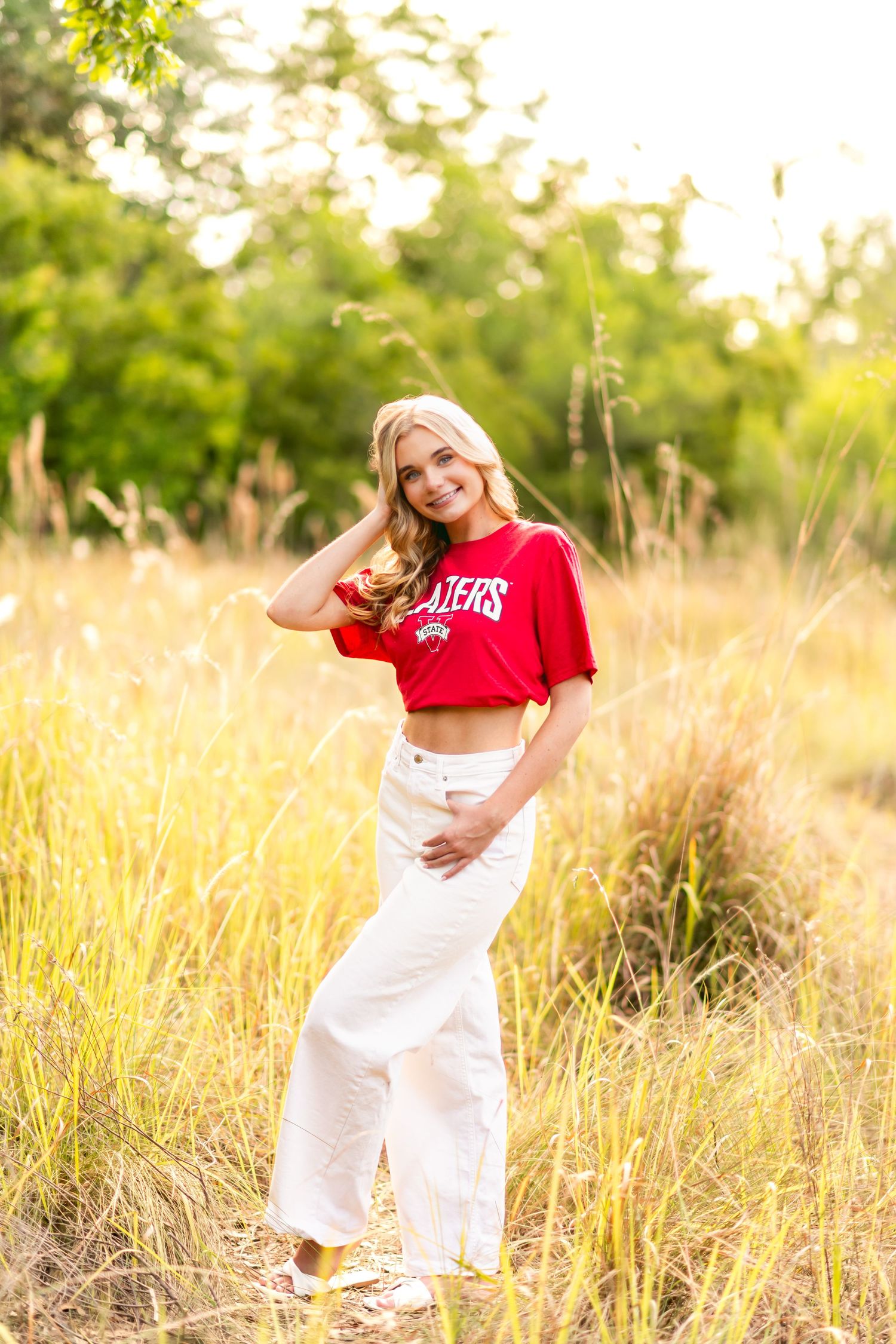 Blonde woman in red crop top and white wide-leg pants posing in golden grass field with green trees background.